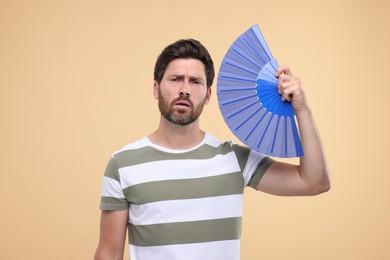Photo of Unhappy man with hand fan suffering from heat on beige background