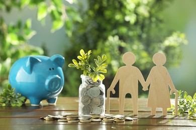 Photo of Pension savings. Figure of senior couple, piggy bank, coins and green twigs on wooden table outdoors