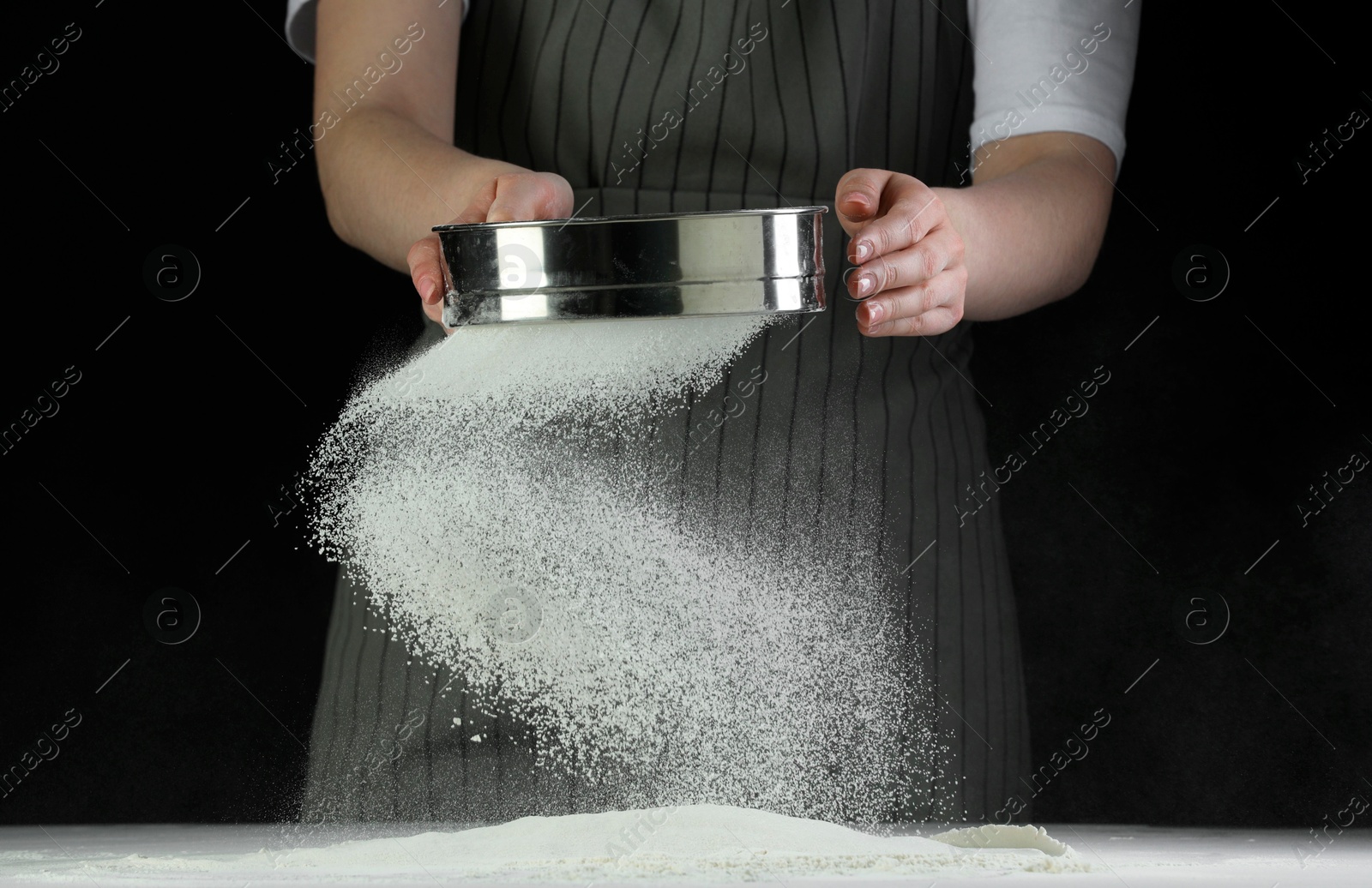 Photo of Woman sieving flour at table against black background, closeup