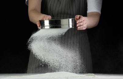 Photo of Woman sieving flour at table against black background, closeup