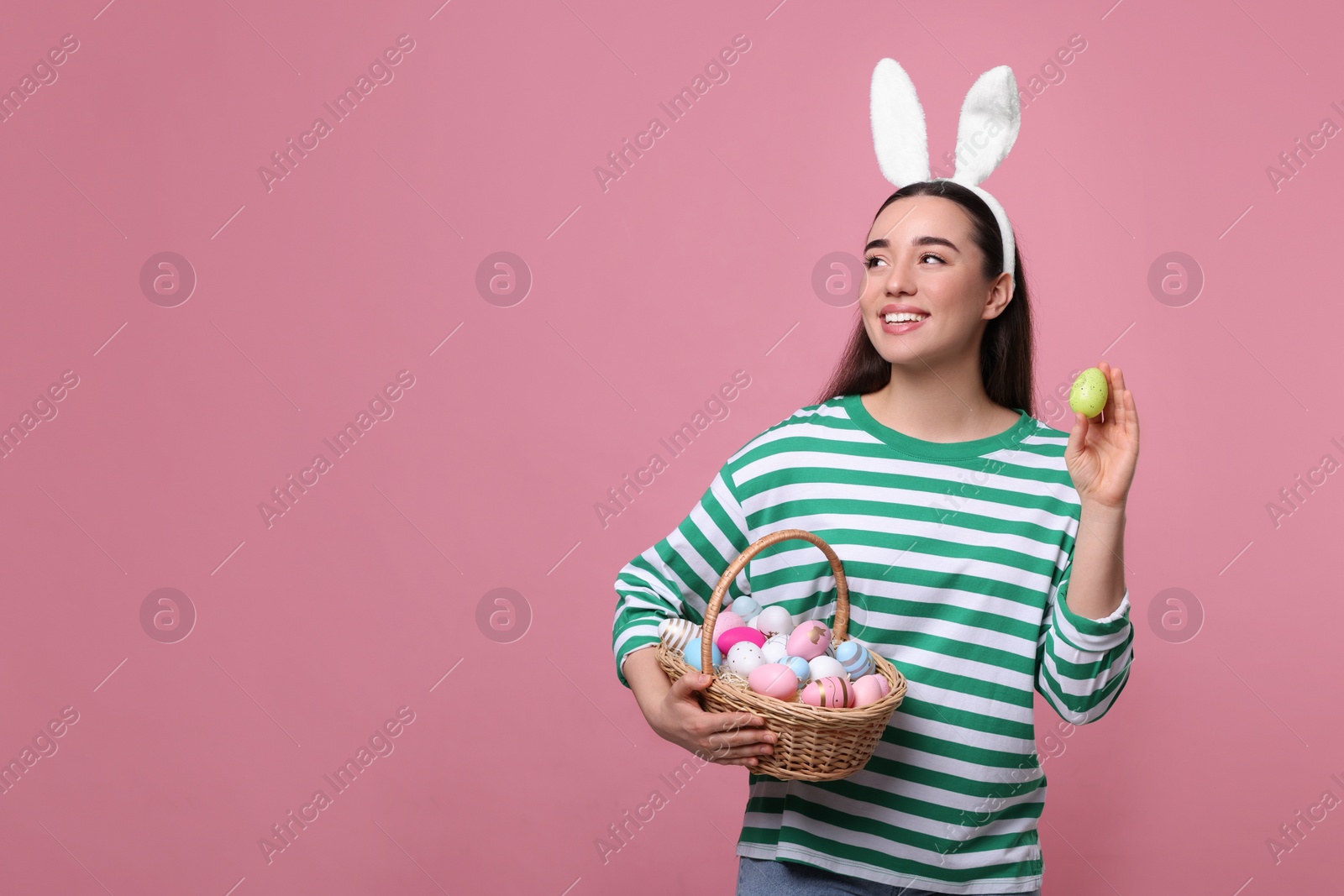 Photo of Happy woman in bunny ears headband holding wicker basket of painted Easter eggs on pink background. Space for text