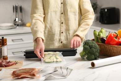 Photo of Woman using sealer for vacuum packing with plastic bag of zucchini at white table in kitchen, closeup
