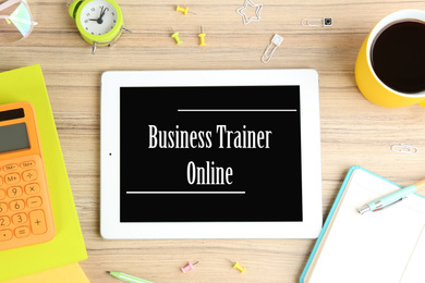 Image of Modern tablet with text BUSINESS TRAINER ONLINE on wooden background, flat lay