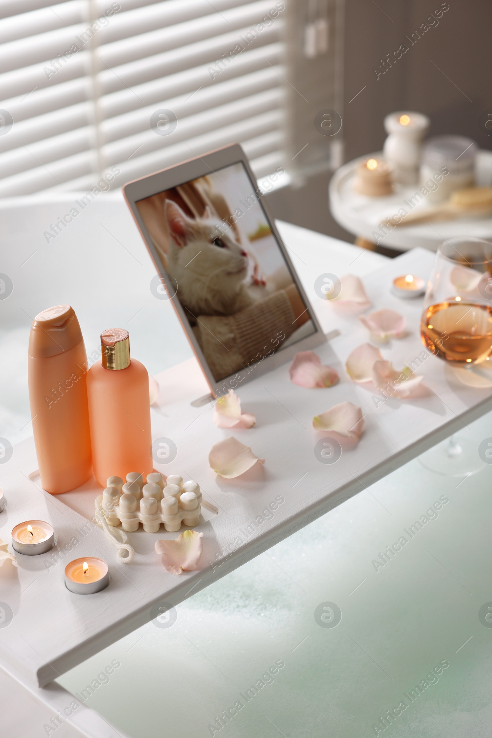 Photo of Wooden tray with tablet, wine, toiletries and flower petals on bathtub in bathroom