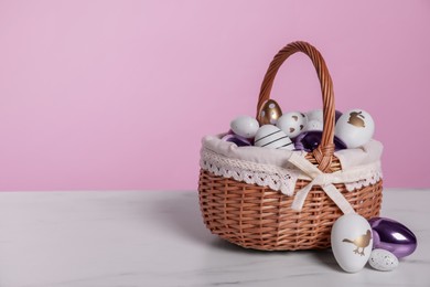 Photo of Wicker basket with festively decorated Easter eggs on white marble table against pink background. Space for text