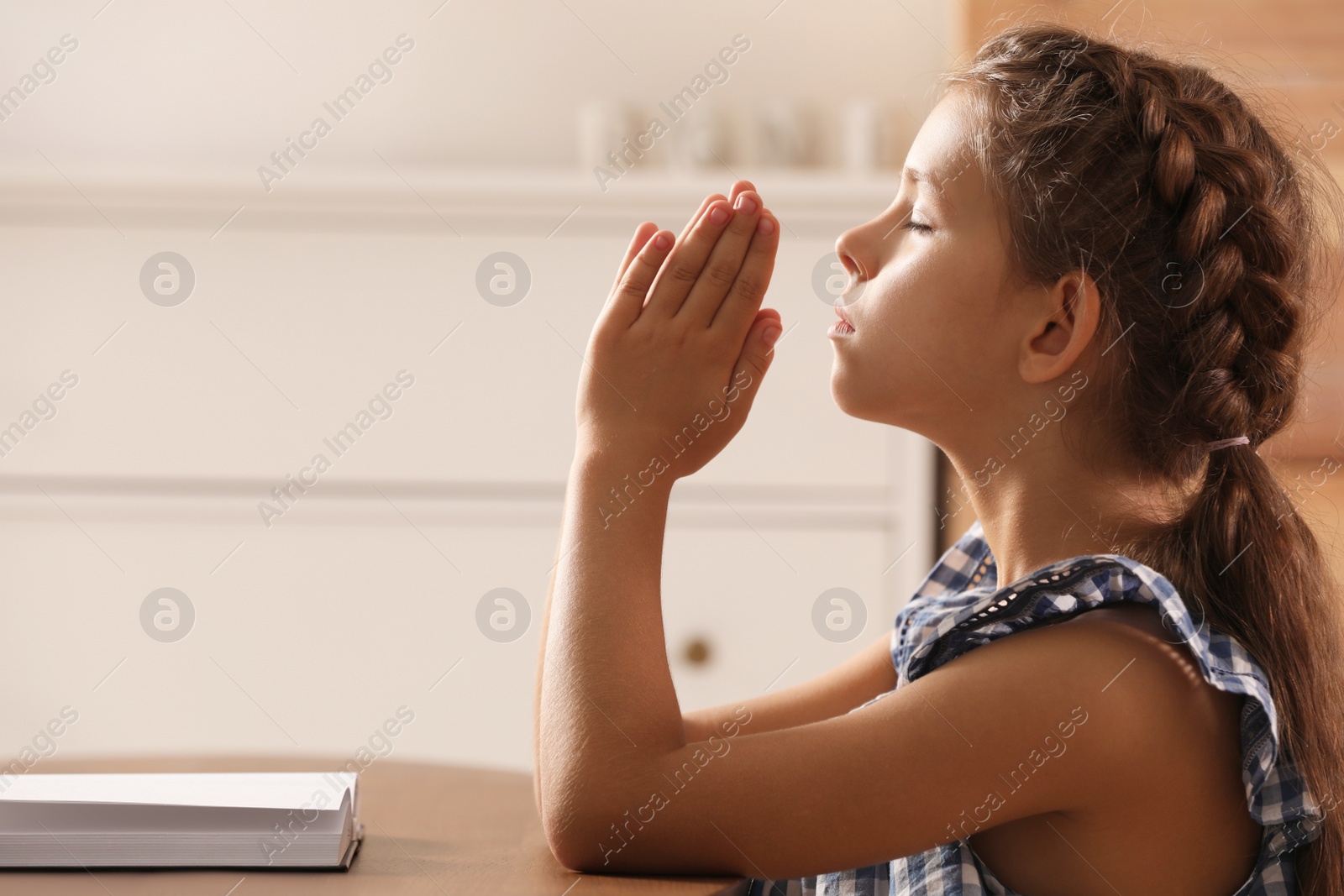 Photo of Cute little girl praying over Bible at table in room