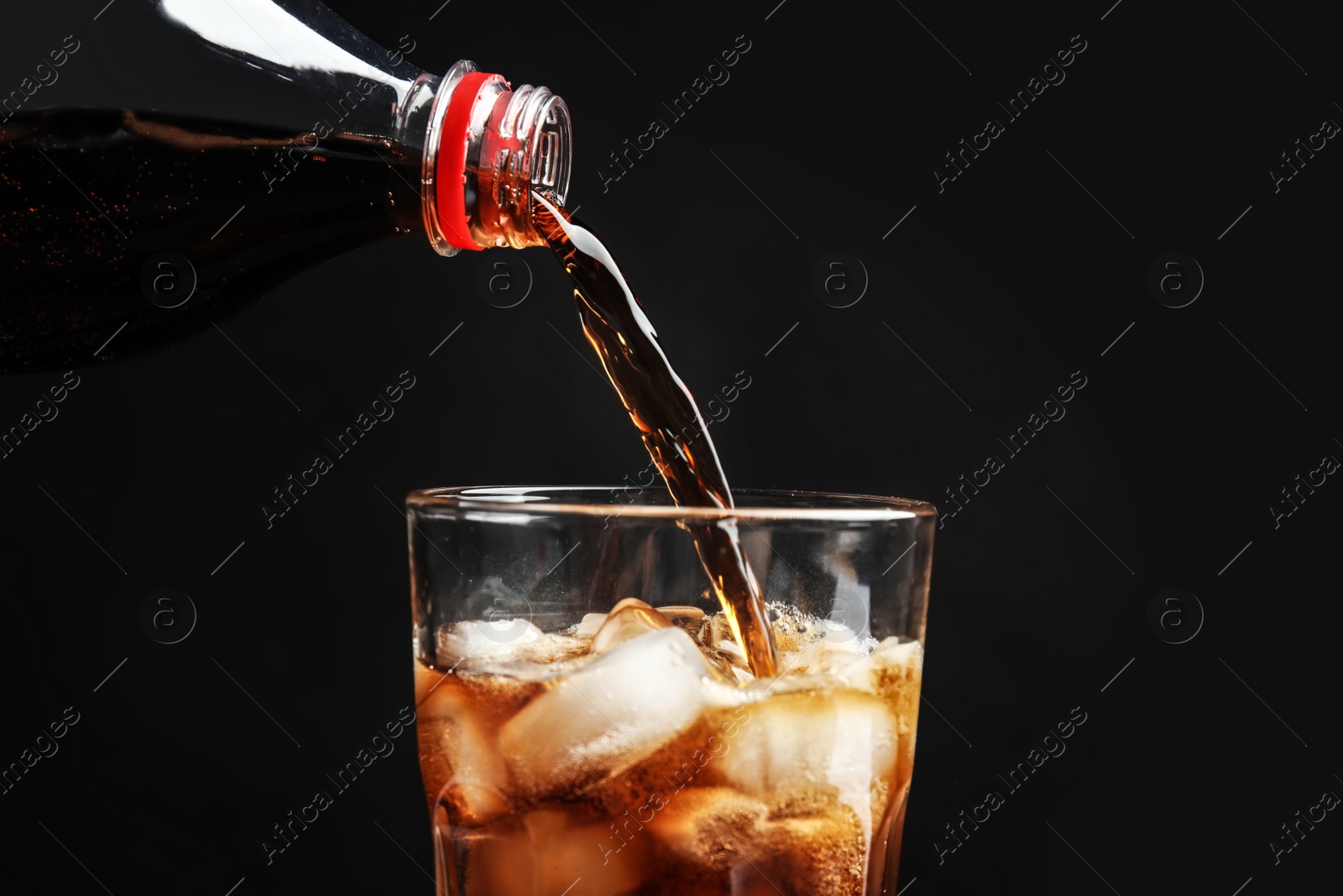 Photo of Pouring refreshing cola from bottle into glass with ice cubes on black background, closeup