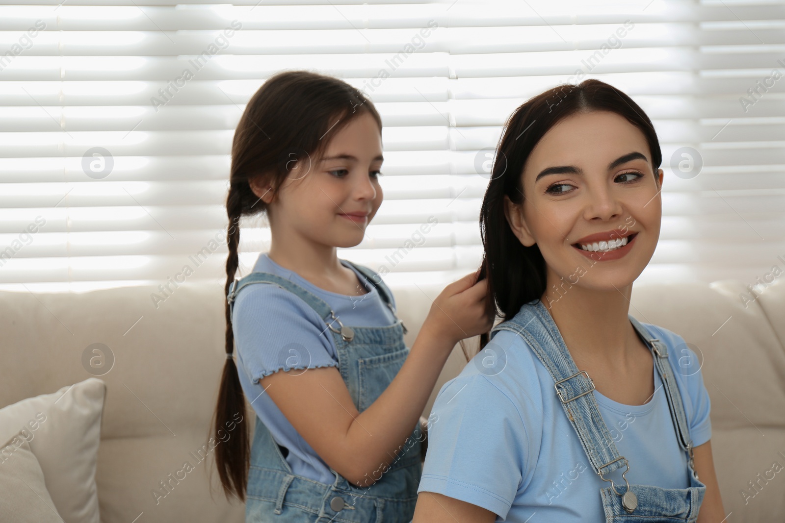 Photo of Cute little girl making mother's hairstyle on sofa indoors