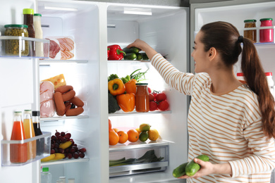 Photo of Young woman taking cucumber out of refrigerator indoors