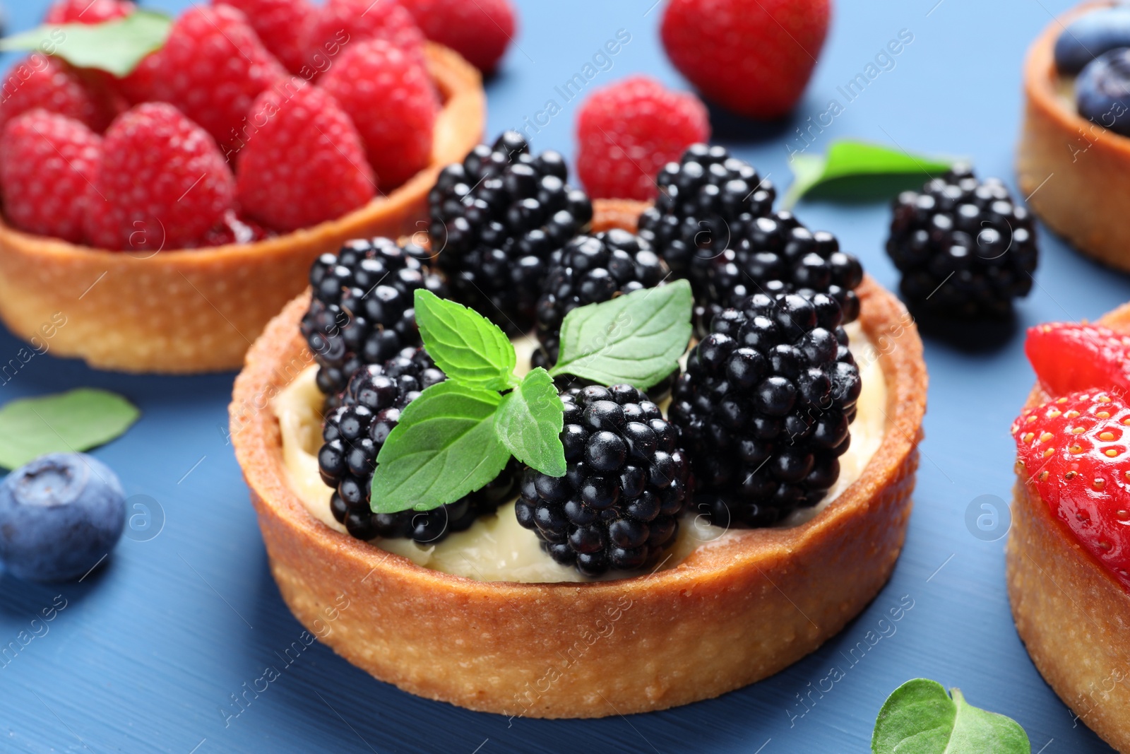 Photo of Tartlets with different fresh berries and mint on blue table, closeup. Delicious dessert