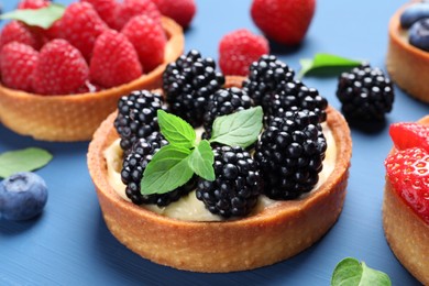 Tartlets with different fresh berries and mint on blue table, closeup. Delicious dessert