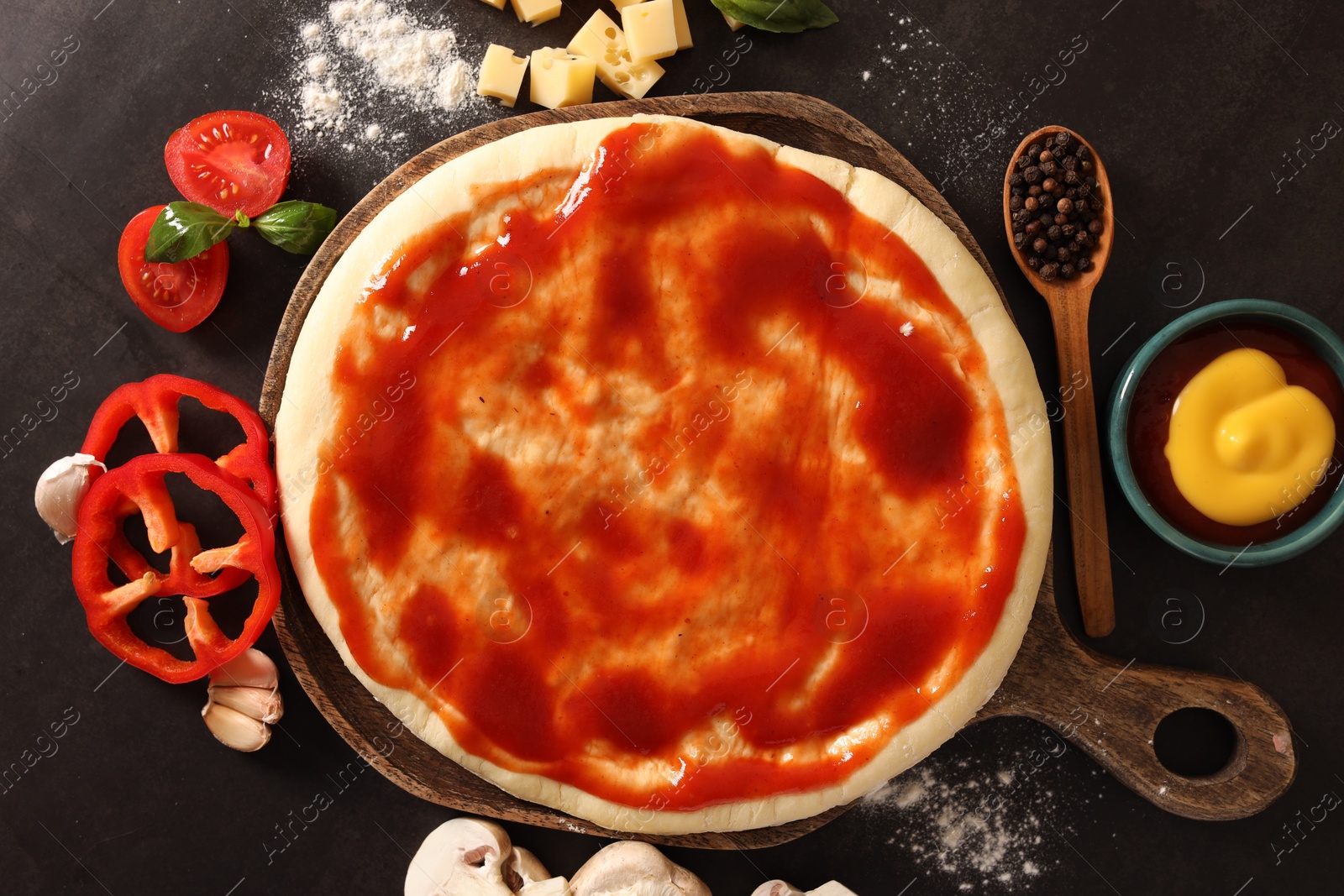 Photo of Pizza dough smeared with tomato sauce and products on dark table, flat lay