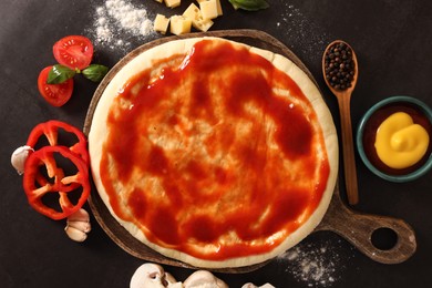 Photo of Pizza dough smeared with tomato sauce and products on dark table, flat lay