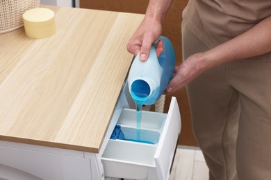 Photo of Man pouring fabric softener from bottle into washing machine indoors, closeup