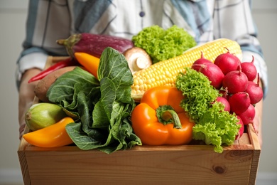 Photo of Woman holding wooden crate full of fresh vegetables on light background, closeup