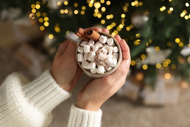 Photo of Woman holding cup of delicious Christmas cocoa with marshmallows and cinnamon sticks against blurred lights, top view