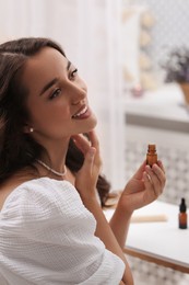 Photo of Beautiful young woman applying essential oil onto neck indoors