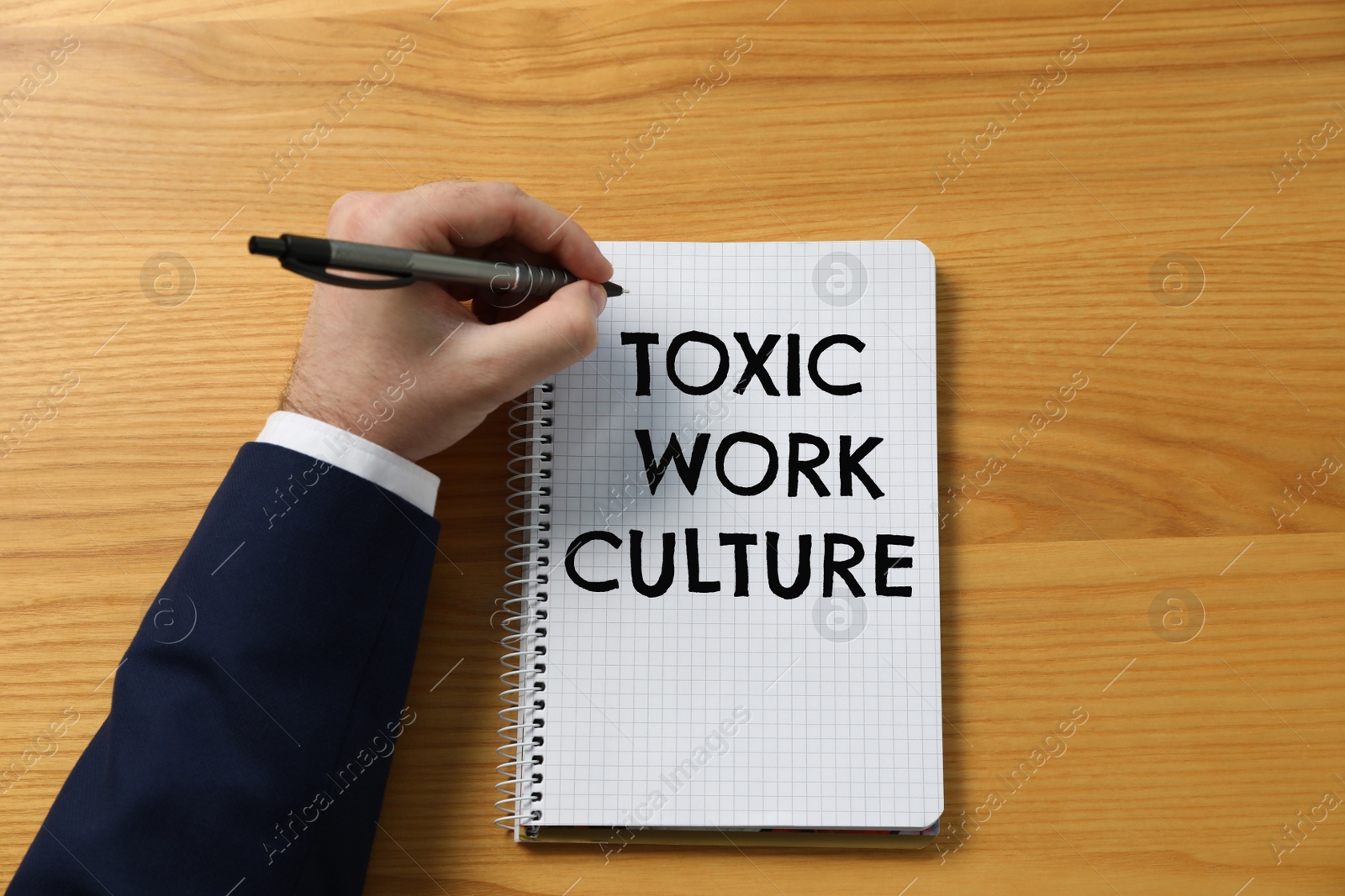 Image of Man writing text Toxic Work Culture in notebook at wooden table, top view