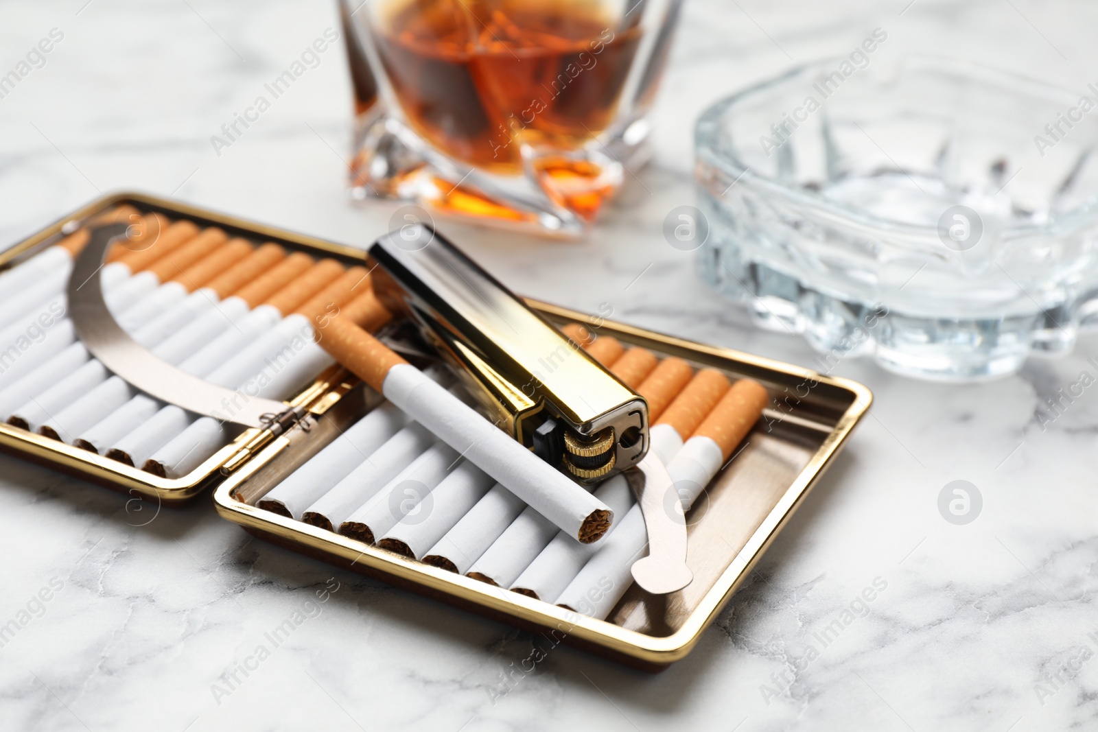 Photo of Open case with tobacco filter cigarettes, lighter, ashtray and alcohol drink on white marble table, closeup