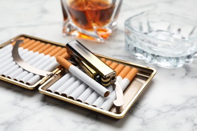 Open case with tobacco filter cigarettes, lighter, ashtray and alcohol drink on white marble table, closeup