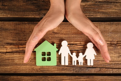 Photo of Woman holding hands near figures of house and family on wooden background, top view