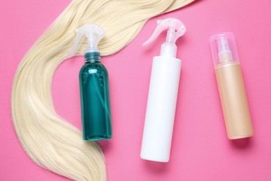 Photo of Spray bottles with thermal protection and lock of blonde hair on pink background, flat lay