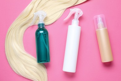 Spray bottles with thermal protection and lock of blonde hair on pink background, flat lay