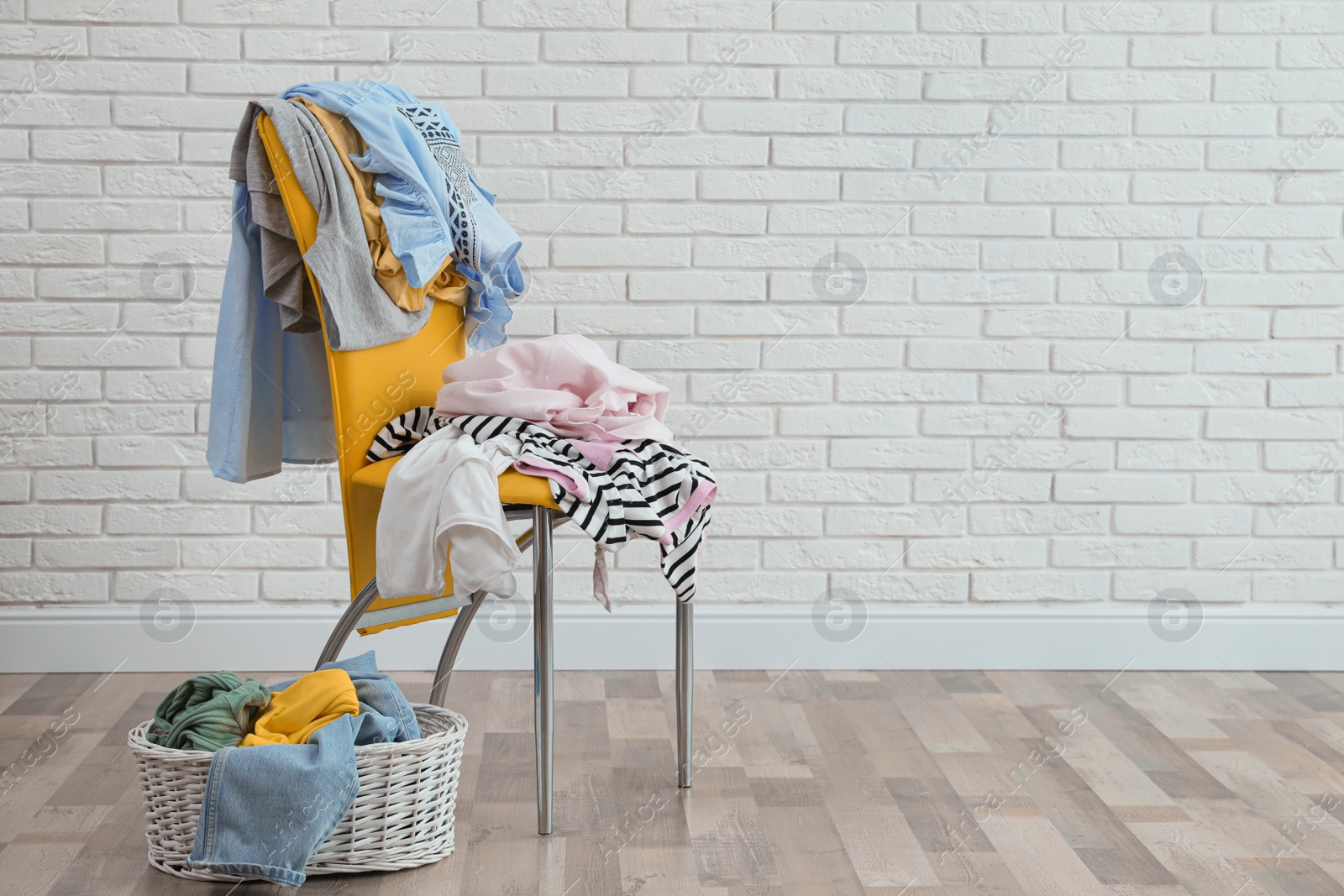 Photo of Yellow chair and wicker basket with different clothes near white brick wall, space for text