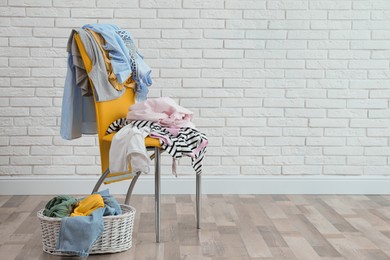 Yellow chair and wicker basket with different clothes near white brick wall, space for text