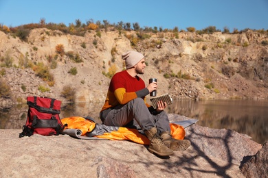 Photo of Male camper with thermos reading book while sitting on sleeping bag in wilderness