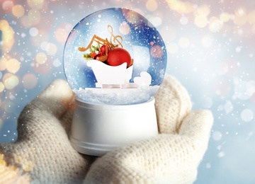 Woman holding snow globe with Christmas decor and music notes on blurred background, closeup. Bokeh effect 