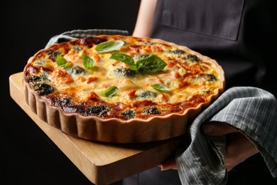 Woman holding wooden board with delicious homemade salmon quiche on black background, closeup