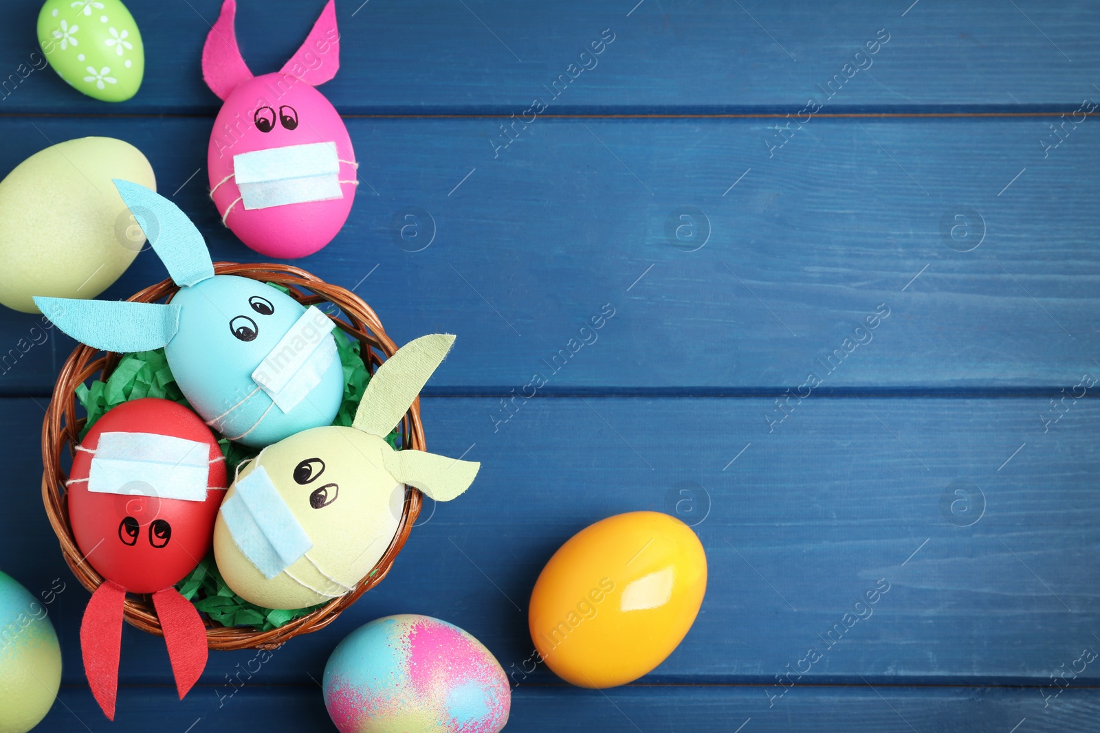 Photo of Painted eggs decorated with bunny ears, protective masks and space for text on blue wooden table, flat lay. Easter holiday during COVID-19 quarantine