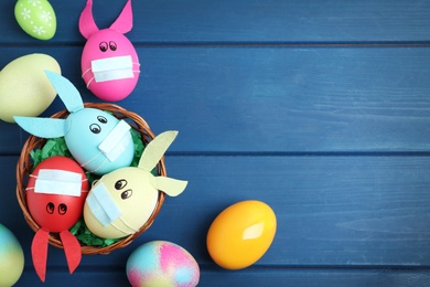 Photo of Painted eggs decorated with bunny ears, protective masks and space for text on blue wooden table, flat lay. Easter holiday during COVID-19 quarantine