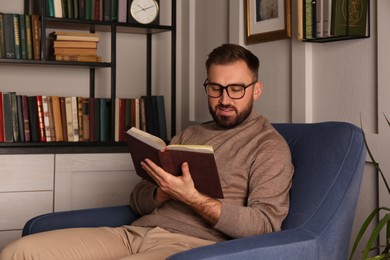 Photo of Young man reading book in armchair indoors. Home library