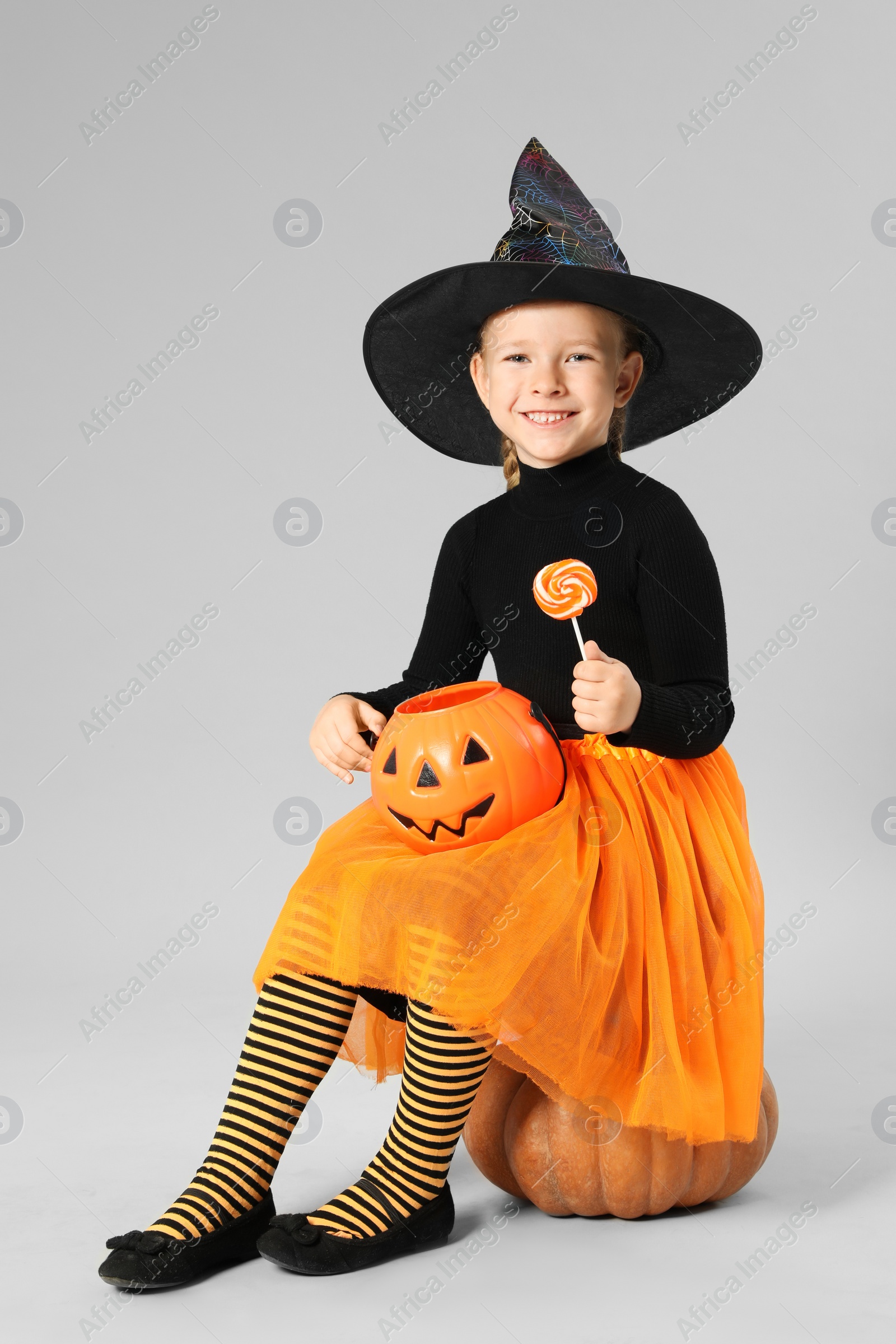 Photo of Cute little girl with pumpkin candy bucket and lollipop wearing Halloween costume on grey background