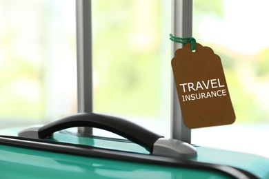 Stylish suitcase with travel insurance label on blurred background, closeup. Space for text