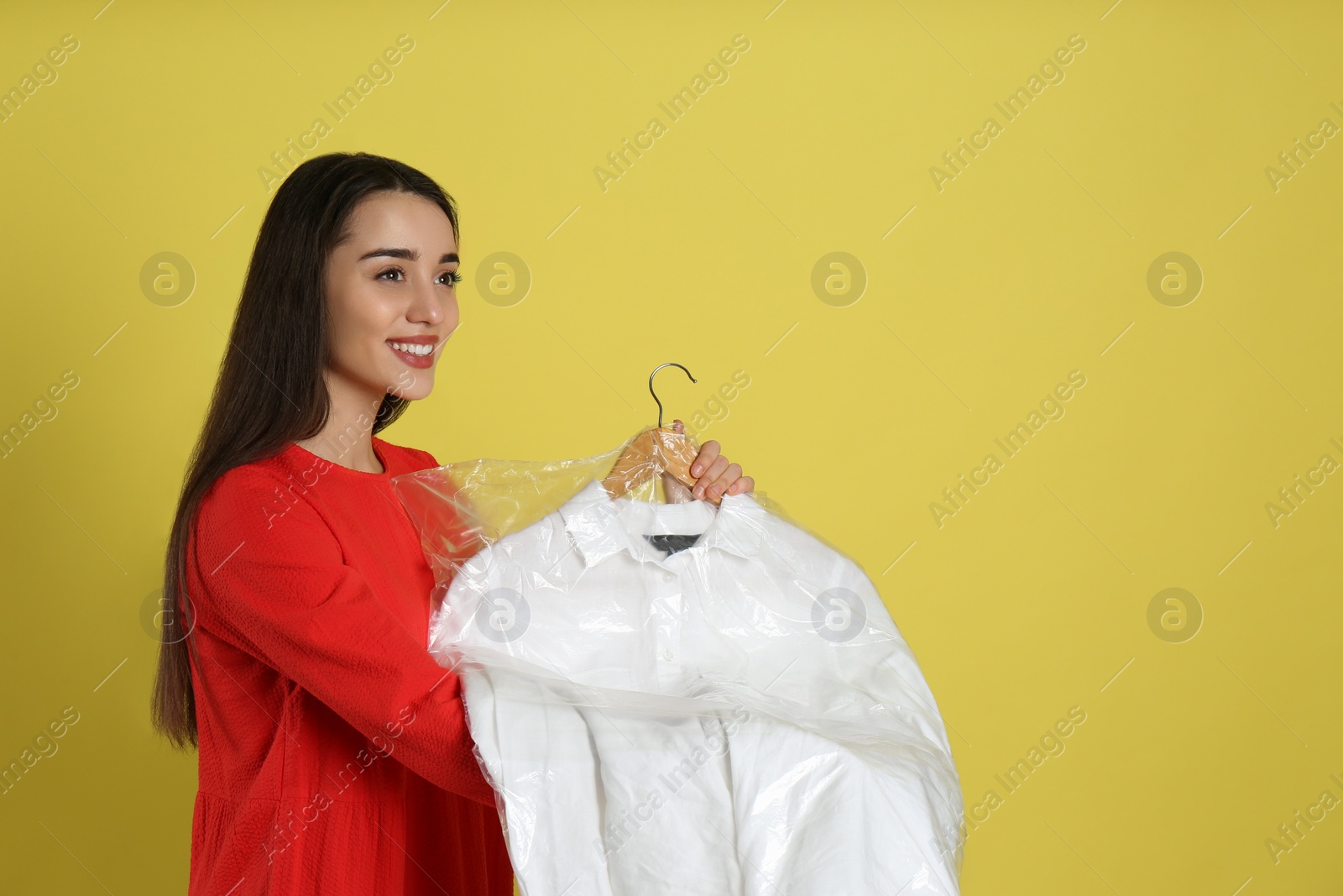 Photo of Young woman holding hanger with shirt in plastic bag on yellow background. Dry-cleaning service