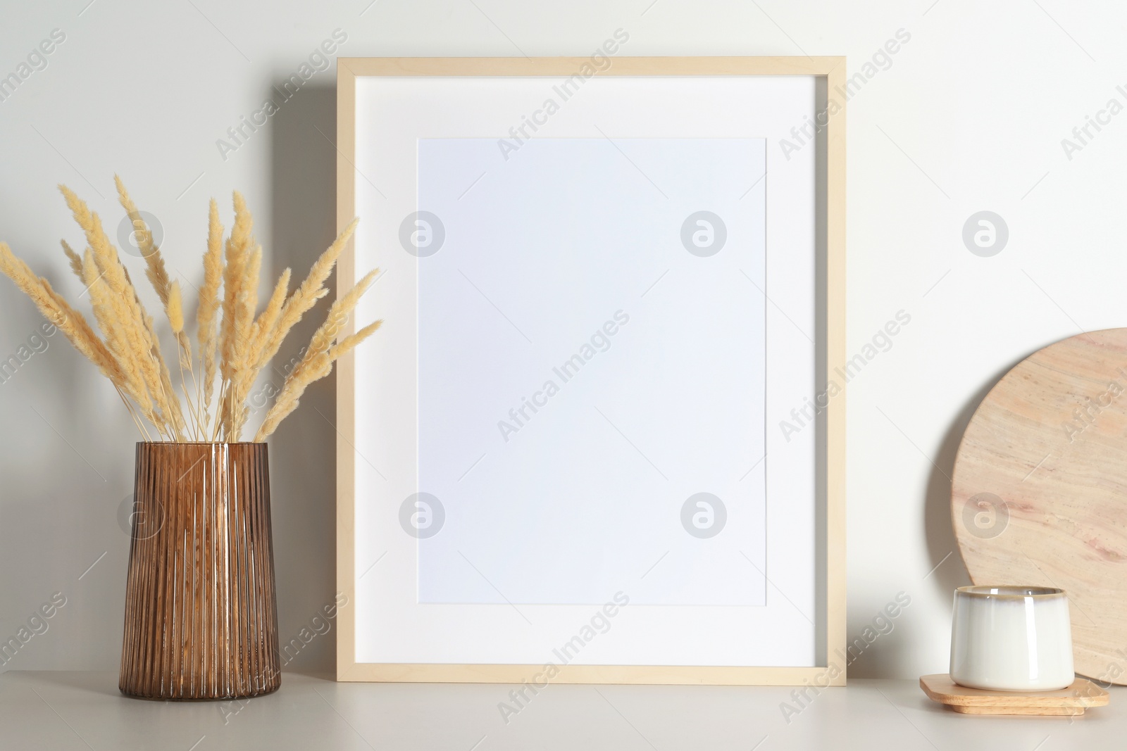 Photo of Empty photo frame, cup and vase with dry decorative spikes on white table. Mockup for design