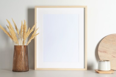 Photo of Empty photo frame, cup and vase with dry decorative spikes on white table. Mockup for design