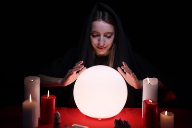 Photo of Soothsayer using glowing crystal ball to predict future at table in darkness. Fortune telling