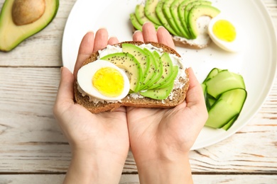 Woman holding crisp toast with avocado, cream cheese and quail egg over table