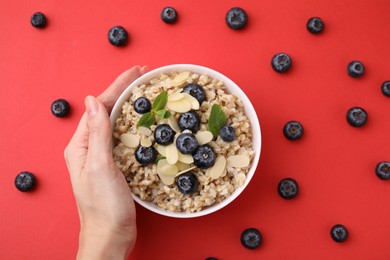 Photo of Woman holding bowl of tasty oatmeal with blueberries, mint and almond petals on red background, top view