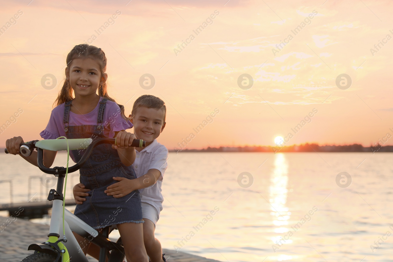 Photo of Happy little siblings riding bicycle near river at sunset