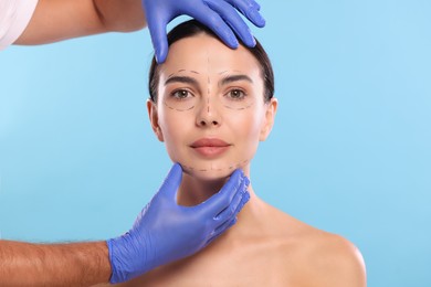 Doctor checking marks on woman's face for cosmetic surgery operation against light blue background