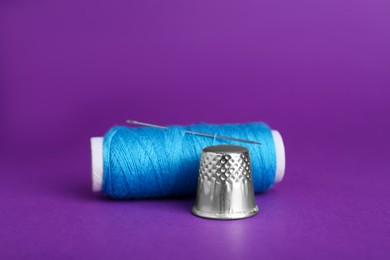 Photo of Thimble and spool of light blue sewing thread with needle on purple background, closeup