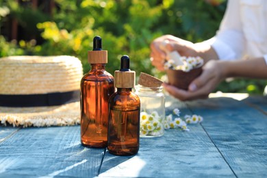 Photo of Woman grinding chamomile flowers in mortar outdoors, focus on bottles of essential oil