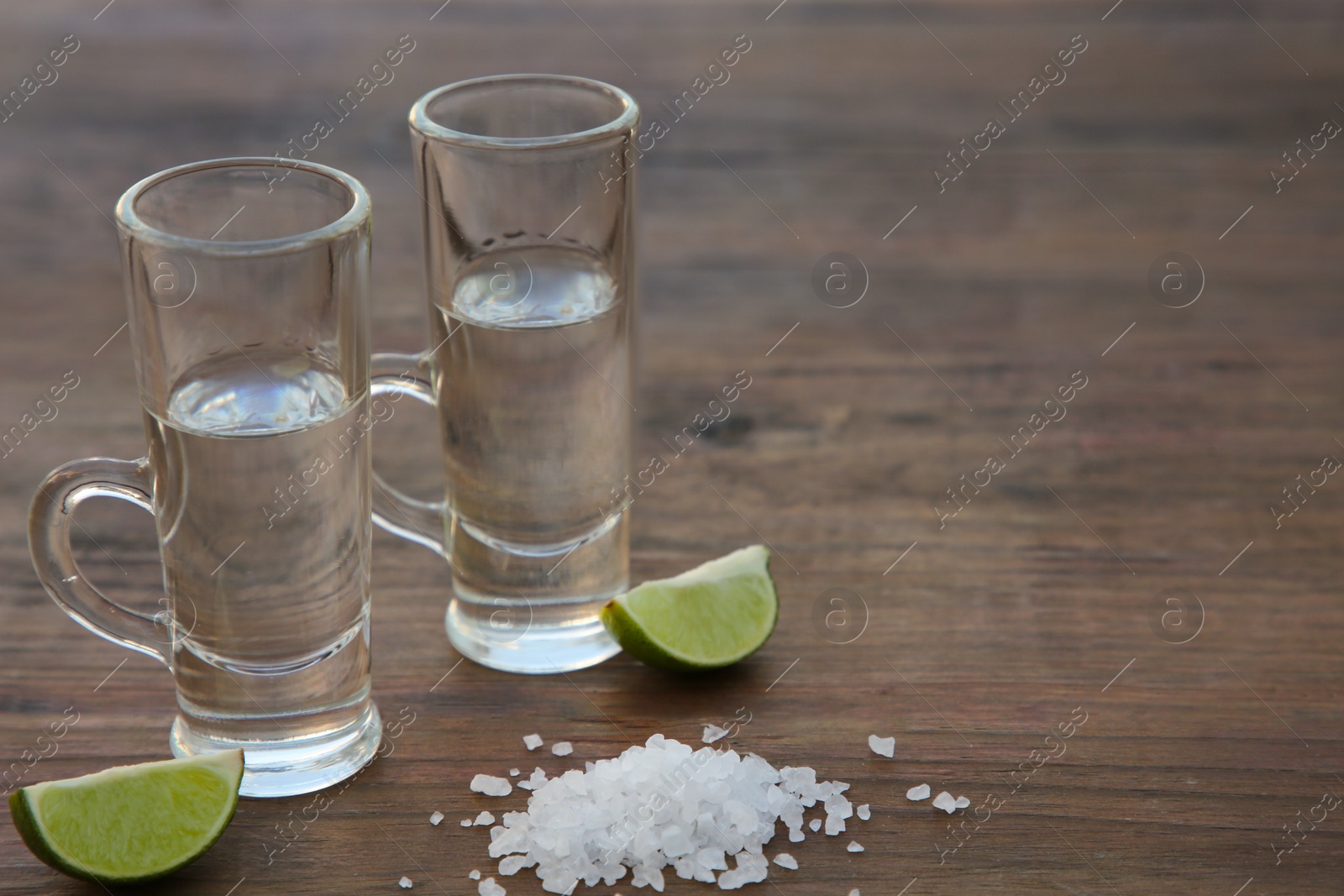 Photo of Mexican tequila shots with lime slices and salt on wooden table, closeup. Space for text