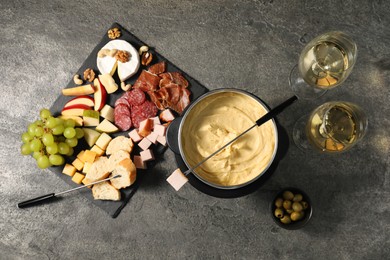 Photo of Fork with piece of ham, fondue pot with melted cheese, wine and snacks on grey textured table, flat lay