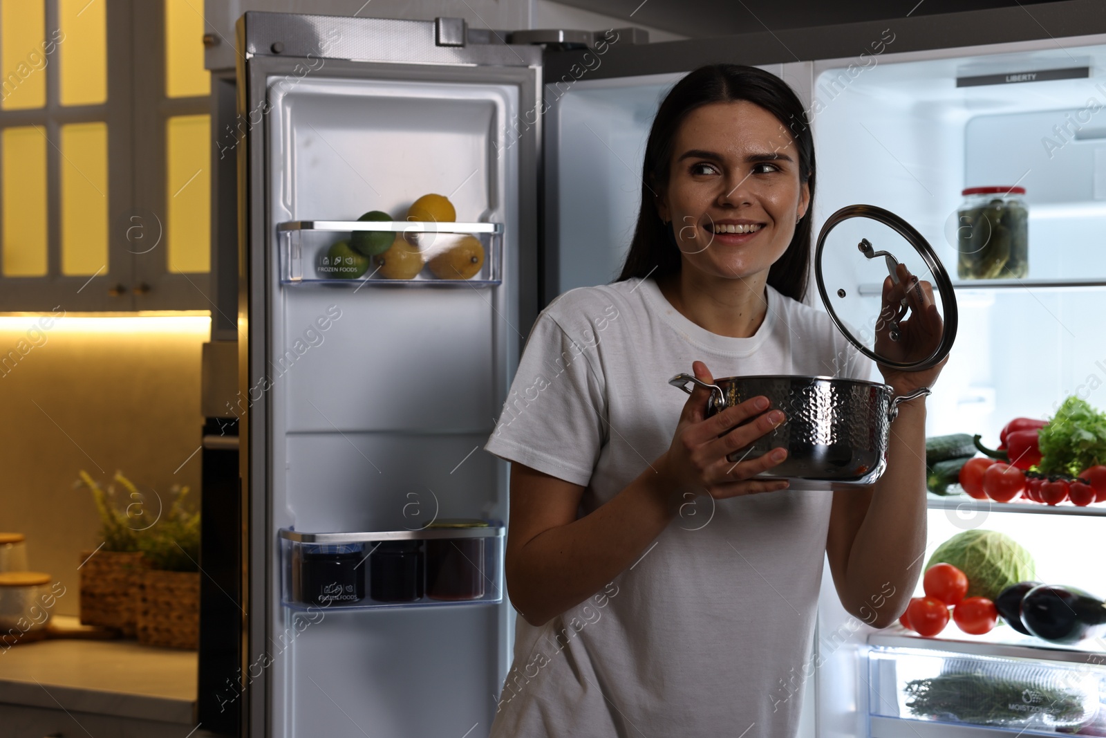 Photo of Young woman with pot near modern refrigerator in kitchen at night